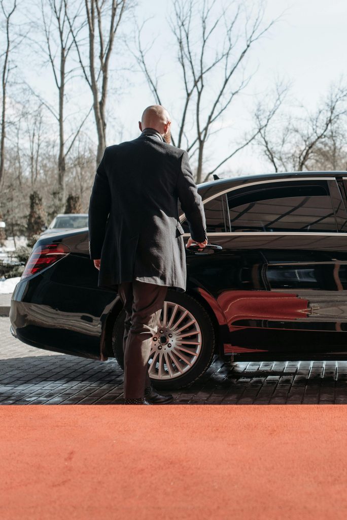 Bald man in a black suit approaches a luxury black car on a sunny day.